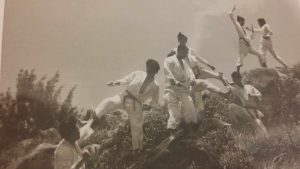 Old sepia tint photo of Taekwon Do students performing various techniques on a hill, in white uniforms with rocks, shrubs and trees behind and around them.
